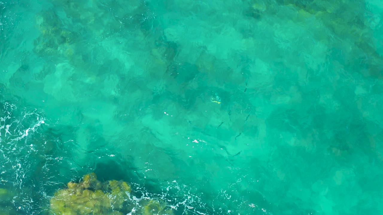 Overhead East Cape water view with blue-green shallows, a rocky patch near the bottom edge, scattered white foam, and no visible people.
