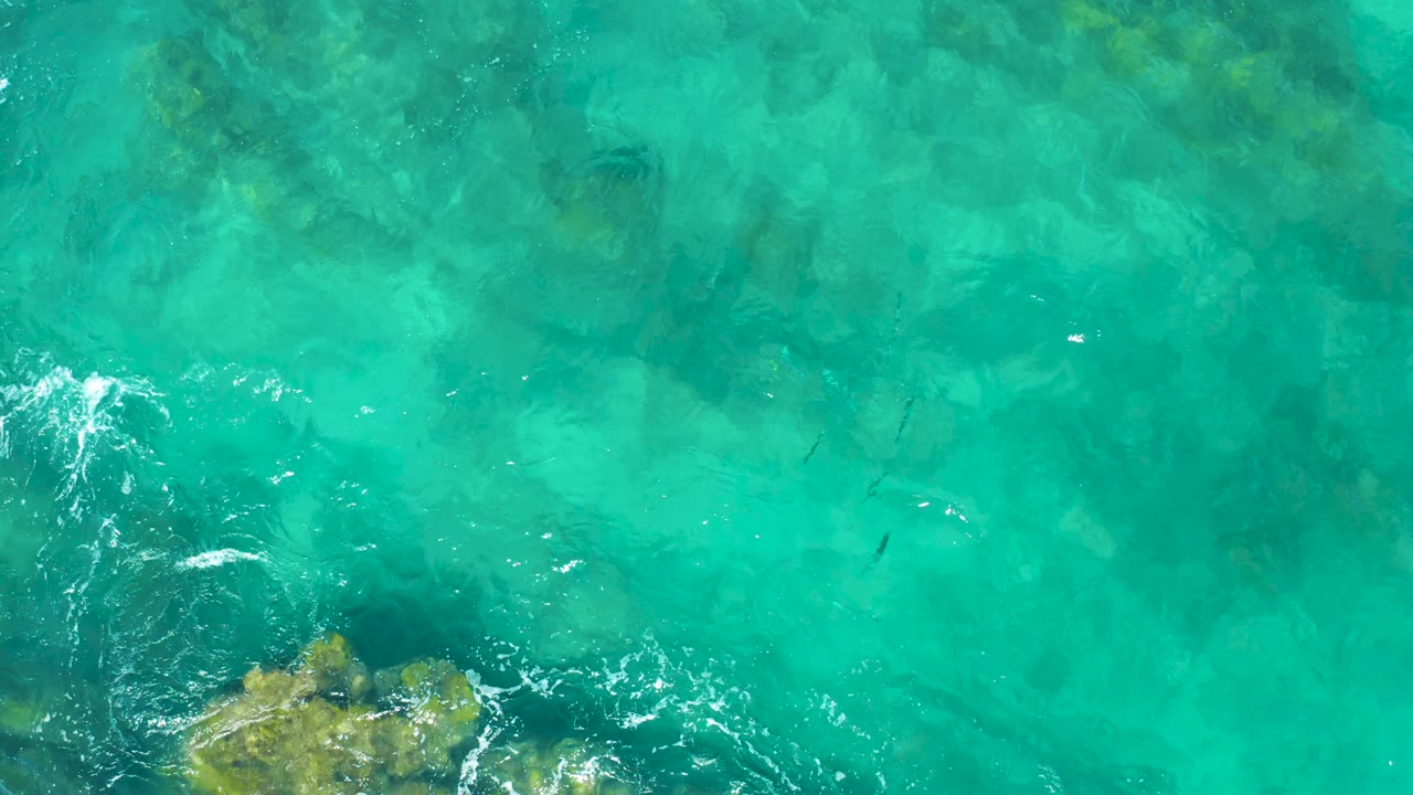 Top-down view of East Cape turquoise shallows with darker underwater rock shapes, light surface ripples, and no visible people.