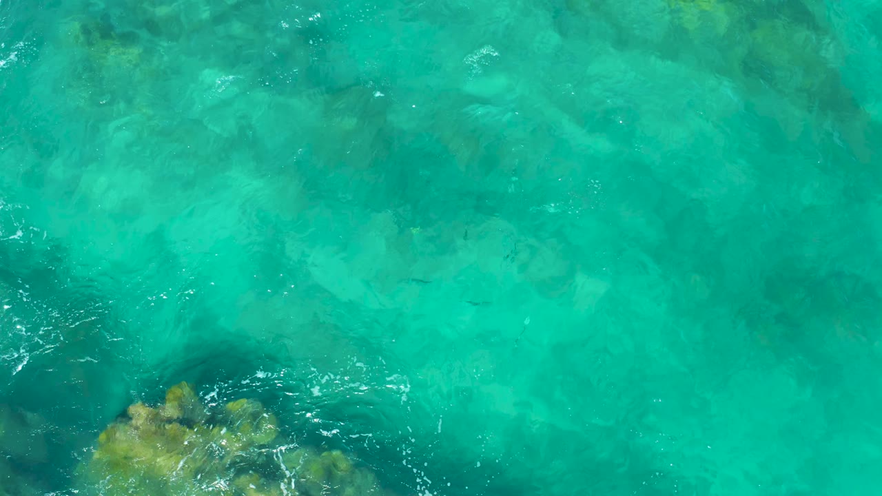 Aerial frame of East Cape turquoise water with pale seabed patterns, small ripple highlights, and a rock cluster partly visible along the bottom edge, no visible people.
