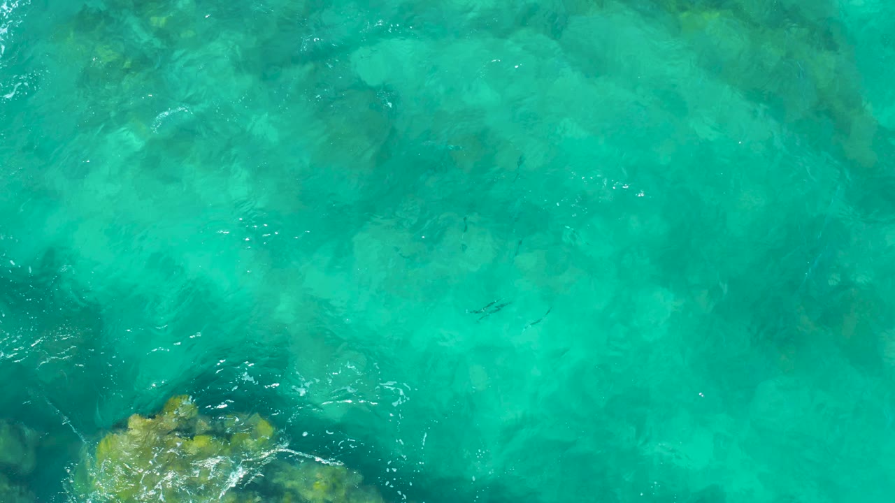 Overhead frame from East Cape showing clear turquoise water, faint underwater stone shapes, a darker rock area at lower left, and no visible people.