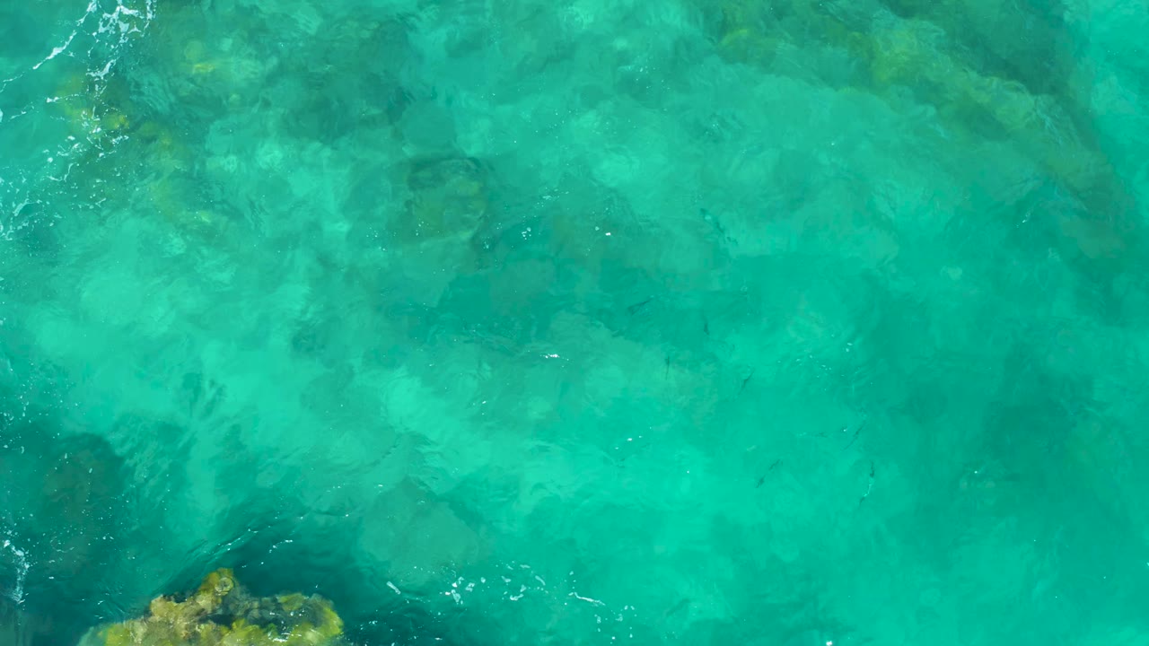 Clear teal ocean water in East Cape, Baja California Sur, shows mottled underwater rock shapes and thin white foam streaks near the top, with no visible people.
