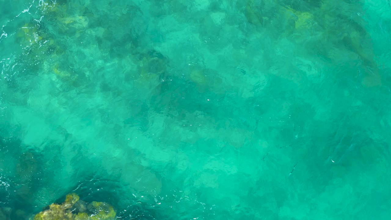 Turquoise water in East Cape, Baja California Sur, fills the frame with dark submerged rock patches and faint surface texture, with no visible people.