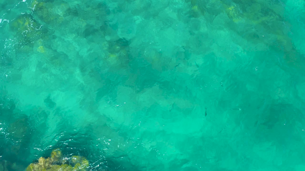 A top-down frame of East Cape, Baja California Sur water shows a yellow-brown rocky outcrop at the lower edge beneath clear turquoise waves, with no visible people.