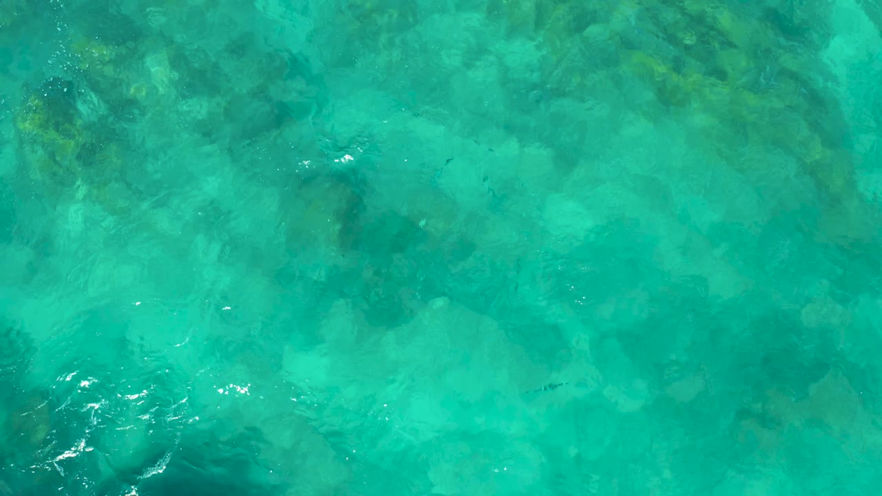 Clear green-blue ocean water in East Cape, Baja California Sur, Mexico, shows submerged rocks, faint fish shapes, bright surface glints, and no visible people.