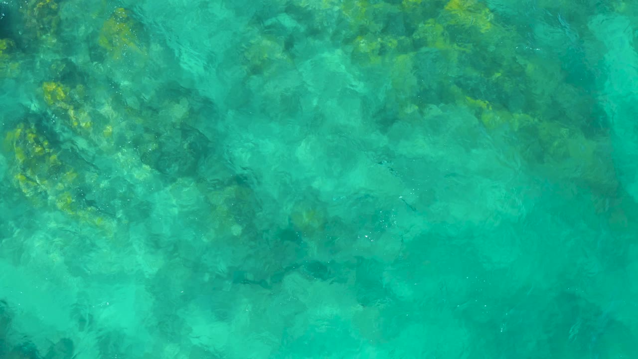 Clear green-blue ocean water in East Cape BCS Mexico reveals mottled rocks and coral-like seabed texture beneath small ripples, no visible people.