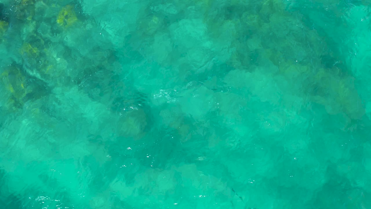 A full-frame overhead view of transparent turquoise ocean in East Cape BCS Mexico shows broad rocky seabed texture softened by surface movement, no visible people.