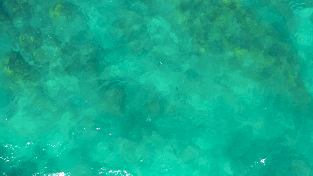 Overhead view of clear turquoise water in East Cape BCS Mexico with darker underwater rock and coral patches visible beneath the rippled surface, no visible people.