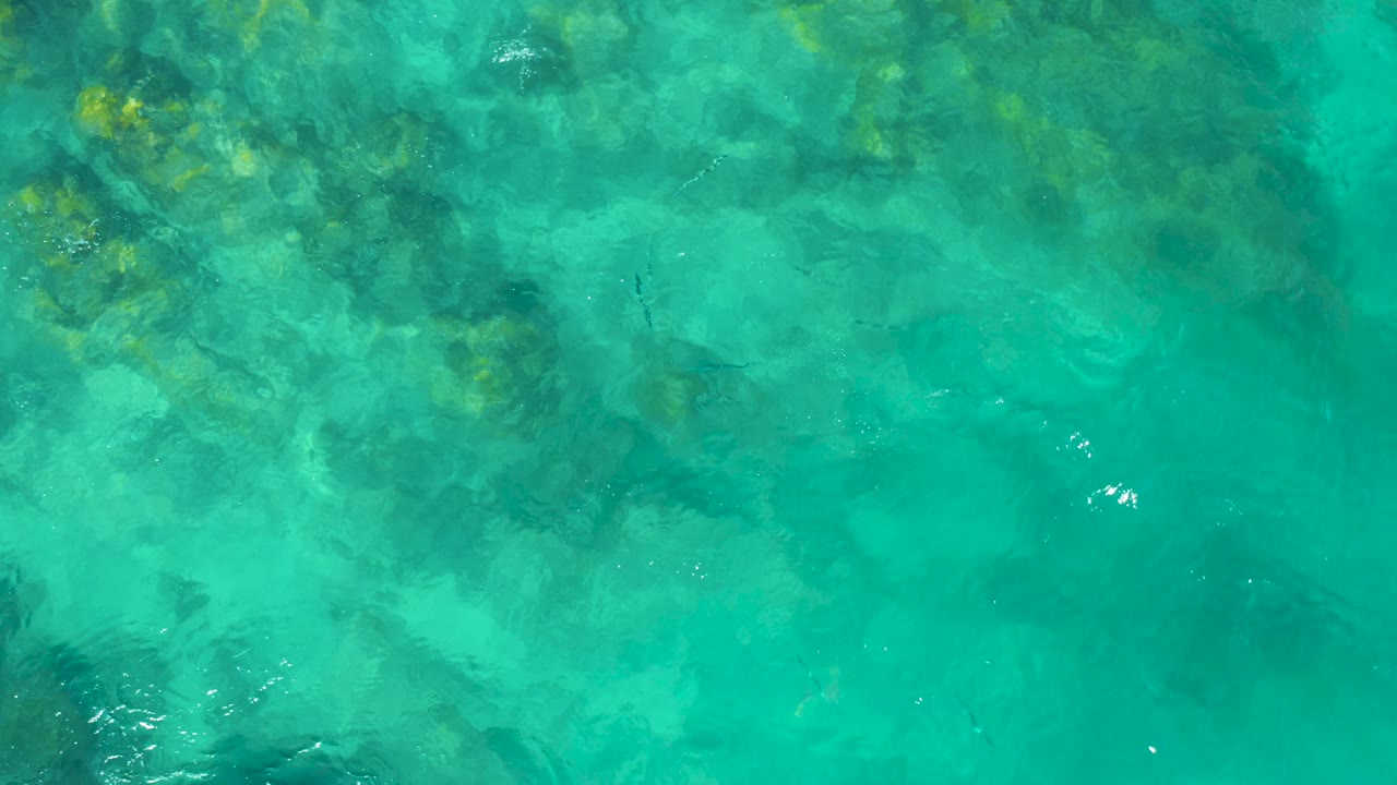 Turquoise shallow water shows green-brown submerged rock and coral areas beneath bright surface flecks in East Cape Baja California Sur, Mexico, with no visible people.
