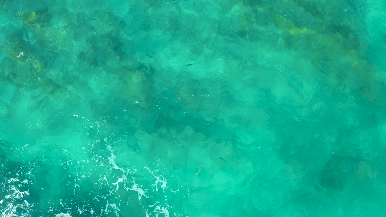 Straight-down aerial view of turquoise East Cape BCS Mexico ocean water with darker seabed patches, small white foam near the lower left, and no visible people.