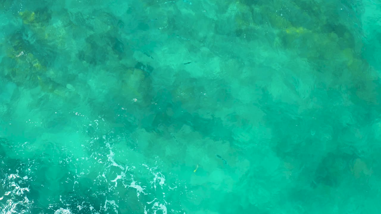 Aerial frame of clear East Cape BCS Mexico water with green-blue surface color, mottled seabed visible below, foam gathering along the lower edge, and no visible people.