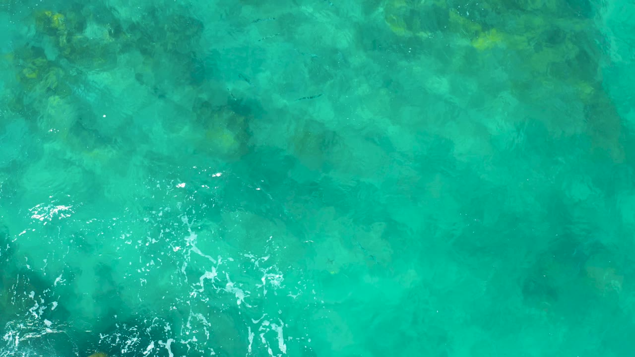 Top-down East Cape BCS Mexico ocean view with translucent turquoise water, darker underwater rock shapes, a small white foam cluster at lower right, and no visible people.