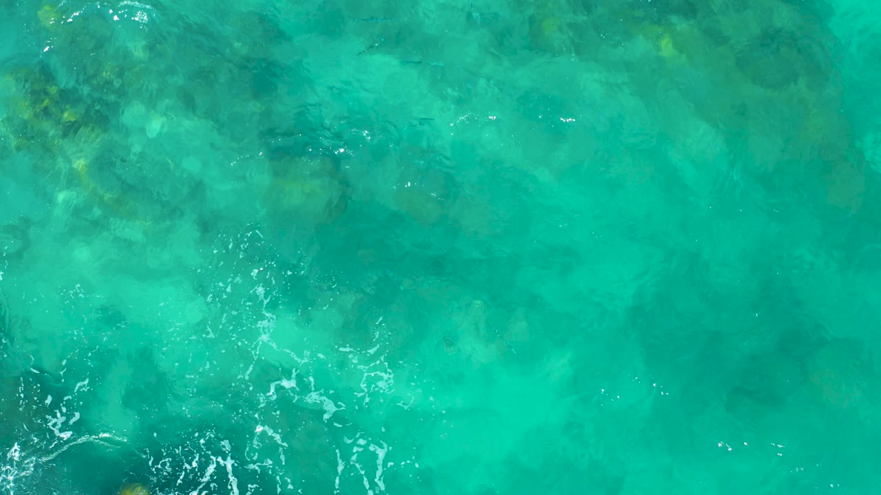 Green-blue ocean water with mottled seabed texture and white foam along the lower left.