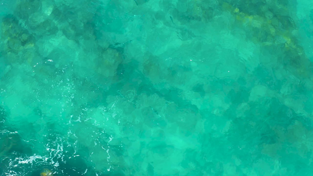 Clear turquoise shallows in East Cape, Baja California Sur, showing mottled underwater rocks and small surface ripples with no visible people.