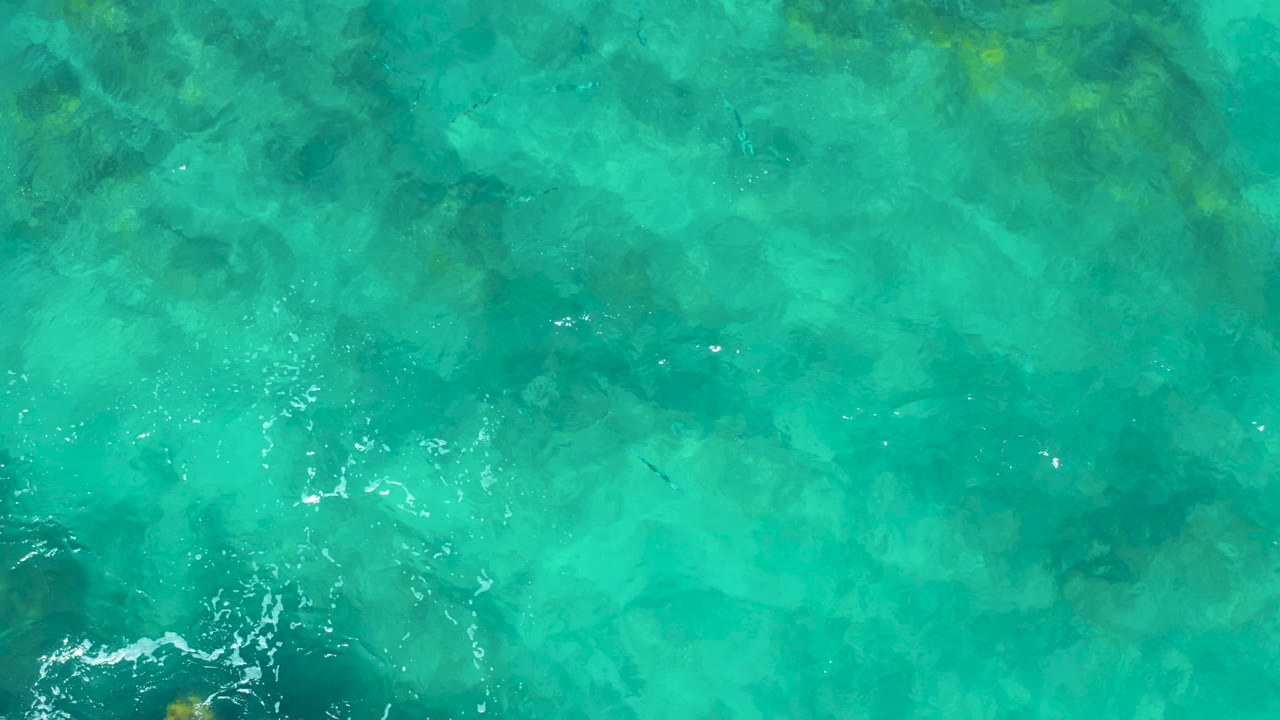 Overhead East Cape water frame with turquoise color, darker submerged formations, and a small cluster of bright surface ripples, with no visible people.