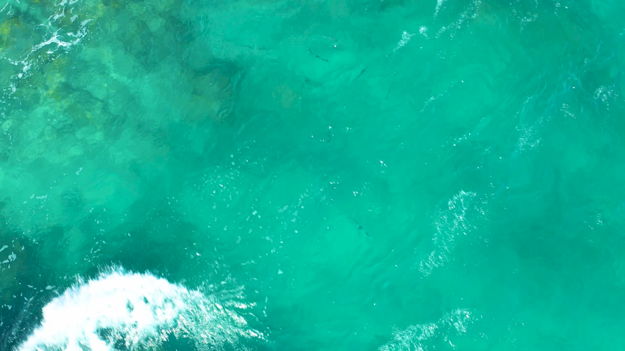 Overhead view of turquoise ocean in East Cape, Baja California Sur, with a white foamy wave at the lower left edge, darker seabed patches beneath the water, and no visible people.