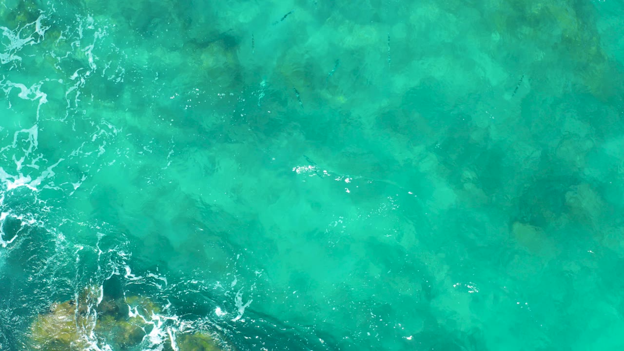 Overhead view of turquoise ocean water in East Cape BCS Mexico with faint submerged rocks, white foam at the left edge, and no visible people.