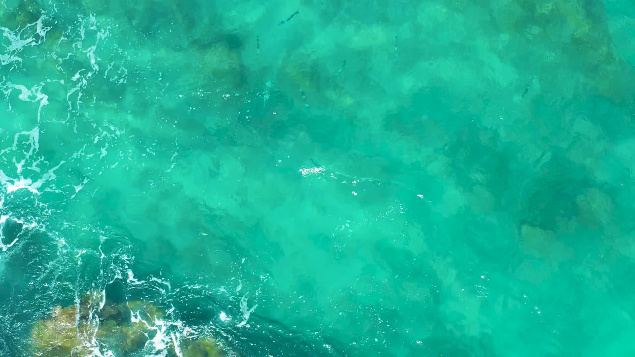 Aerial-looking frame of East Cape BCS Mexico water where white foam drifts over darker submerged rock shapes, with no visible people.