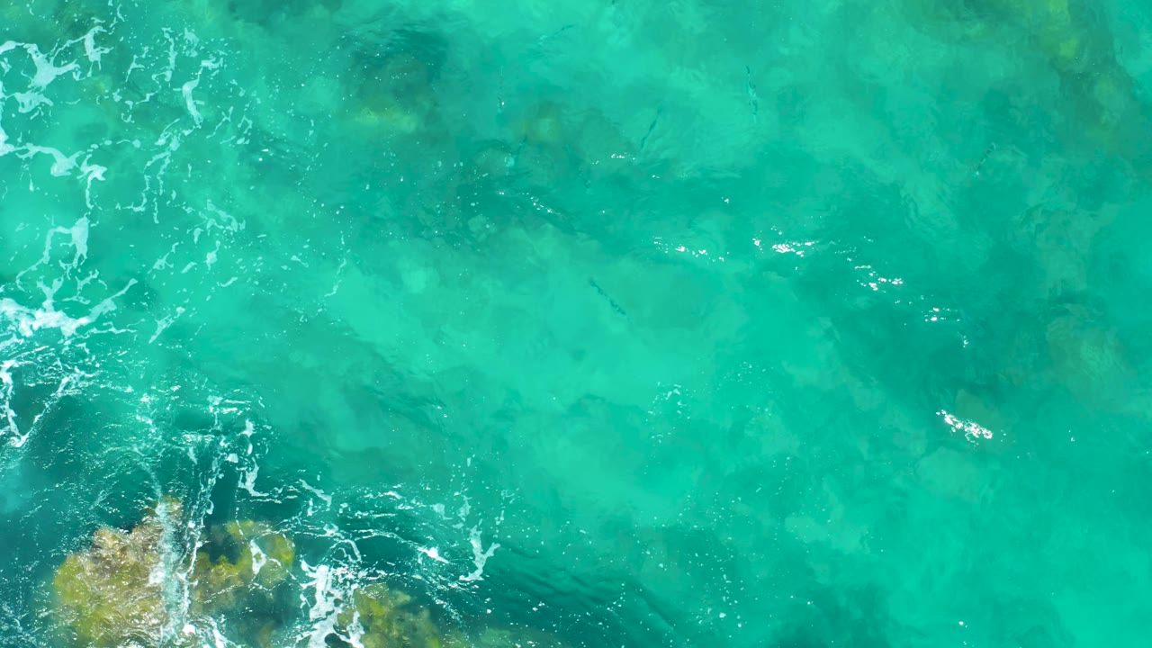 Top-down East Cape BCS Mexico ocean view with a golden-brown underwater rock patch near the lower center, scattered foam, and no visible people.