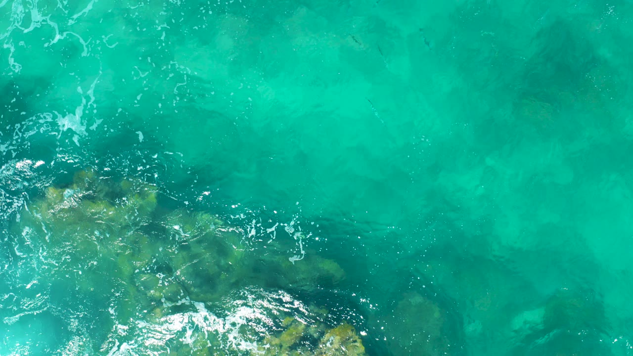 Overhead view of turquoise East Cape ocean water with green-brown submerged rocks near the lower left and no visible people.