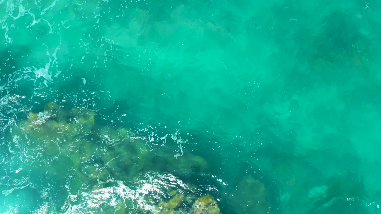 Clear turquoise water in East Cape, Mexico, passes over submerged rock patches along the lower edge, with no visible people.