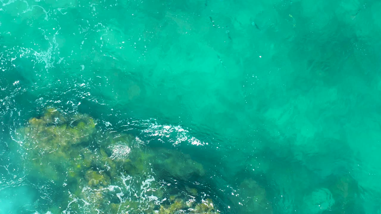 Top-down East Cape ocean frame with underwater rock patches clustered toward the lower center and right, surrounded by clear turquoise water and no visible people.