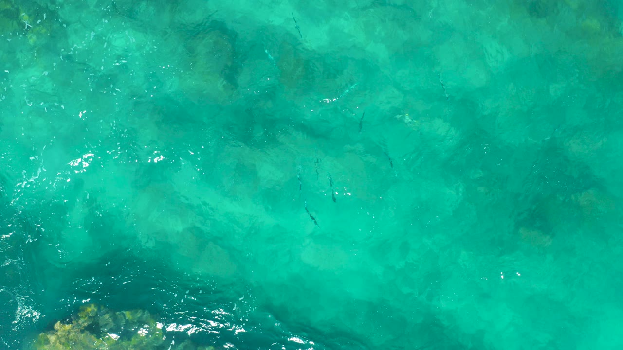 Overhead view of clear turquoise water in East Cape, Baja California Sur, with darker underwater rocks near the lower left and no visible people.