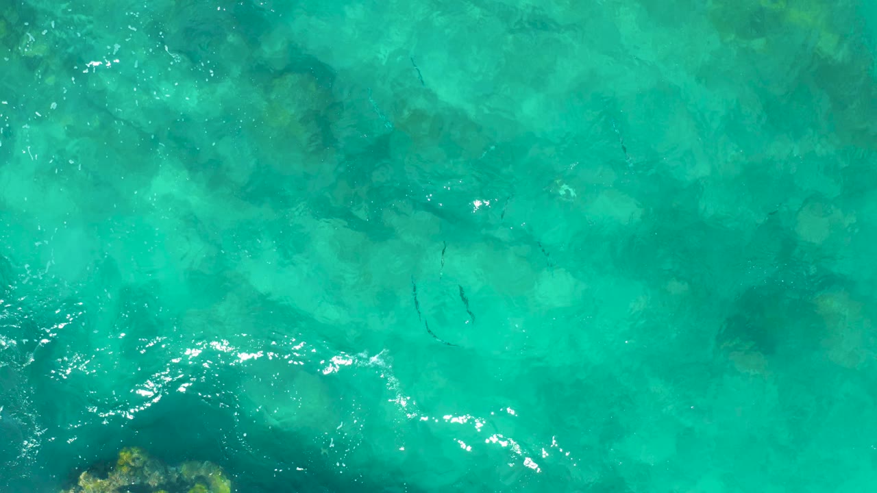 An overhead frame of East Cape shallow water shows bright white reflections across the upper right and darker underwater patches below, with no visible people.