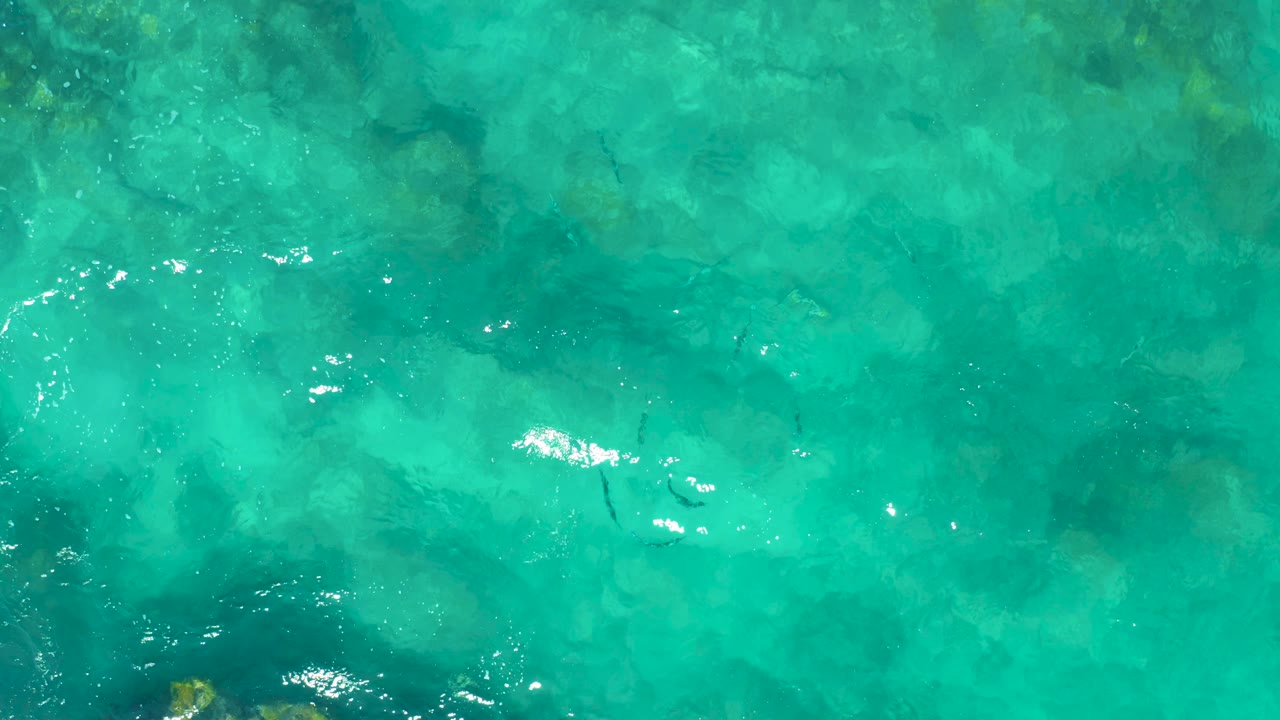 Clear turquoise water in East Cape shows a darker rocky area entering from the lower left and fine surface ripples, with no visible people.