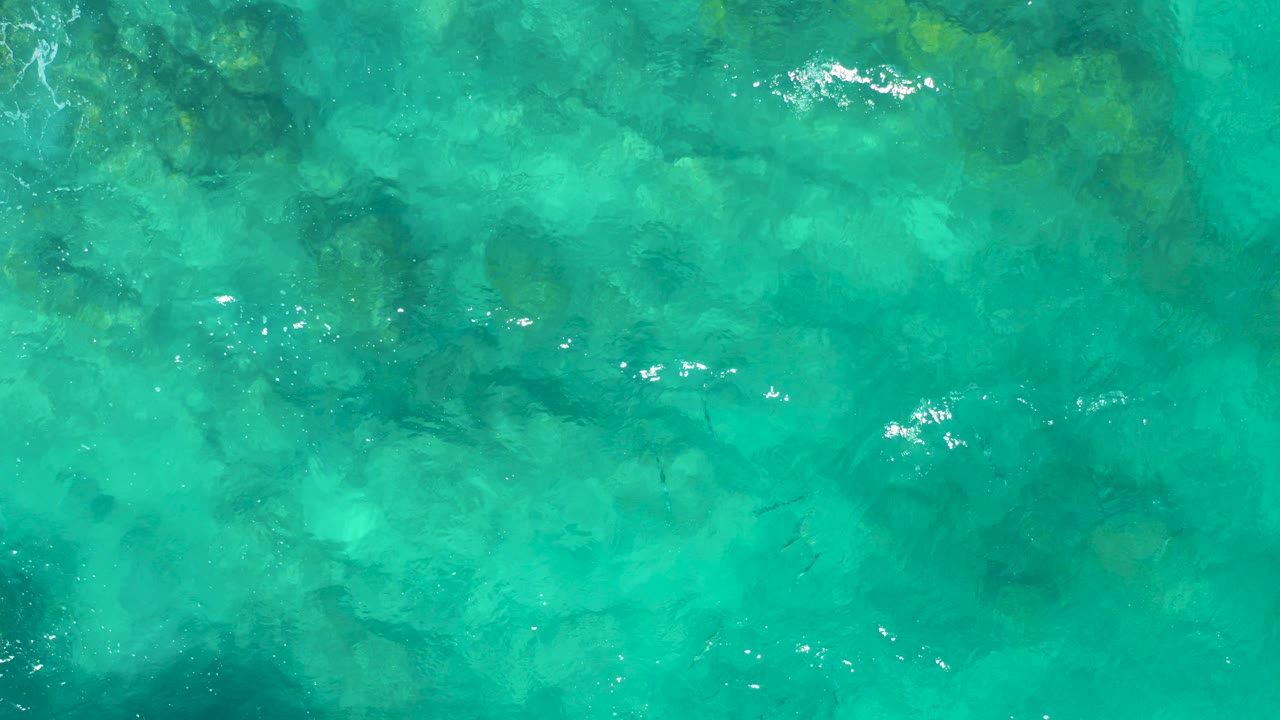 Overhead view of clear turquoise water in East Cape, Baja California Sur, with submerged rocks, faint small fish shapes, surface ripples, and no visible people.