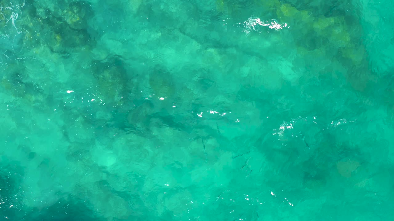 Clear turquoise ocean water in East Cape, Baja California Sur shows darker submerged rock patches, tiny fish silhouettes, light ripples, and no visible people.