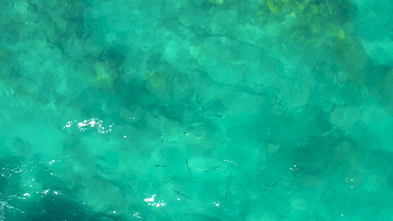 Sunlit turquoise water in East Cape BCS Mexico reveals darker rocky patches, small fish below the surface, bright glints, and no visible people.