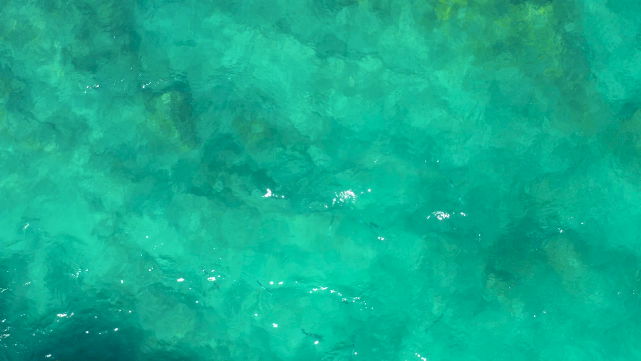 Overhead view of clear turquoise water in East Cape, Baja California Sur, with mottled rocks below the surface, small fish shapes, bright ripple highlights, and no visible people.