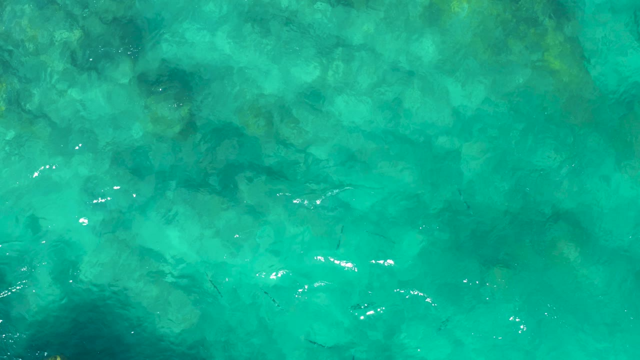 Overhead frame of shallow turquoise water in East Cape, Baja California Sur, where the rocky seabed appears as soft green and blue patches under moving light, with no visible people.