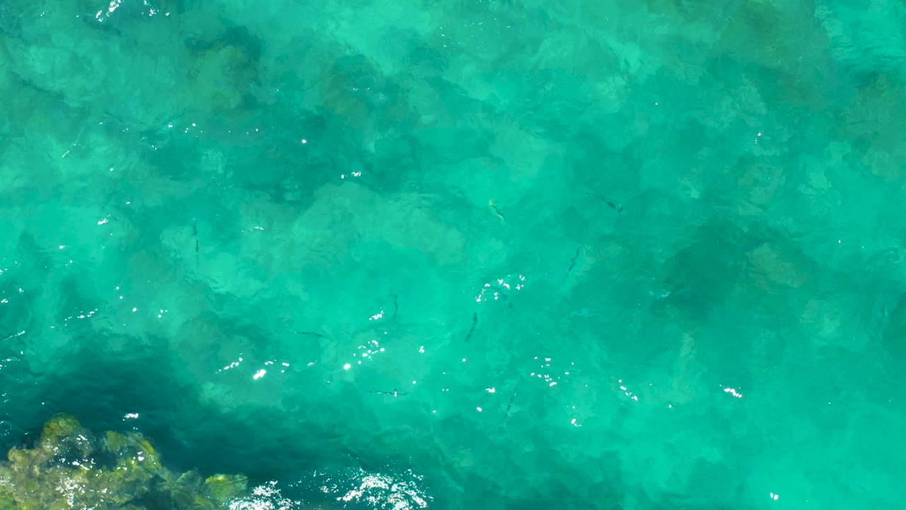 The East Cape BCS Mexico shallows appear bright turquoise with mottled rocks under the surface, a rocky outcrop at the lower left edge, scattered fish shapes, and no visible people.