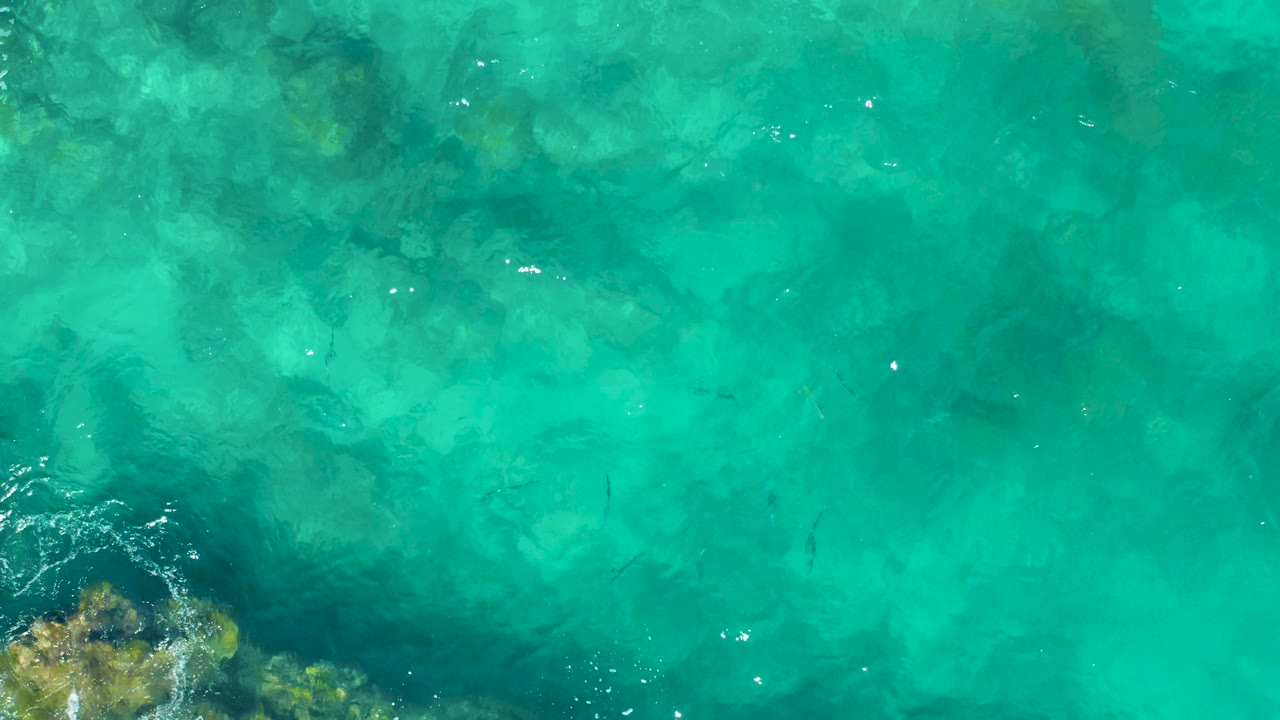 Top-down view over East Cape BCS Mexico shallow ocean, with rocks entering from the bottom, pale seabed patches, small fish marks, ripples, and no visible people.
