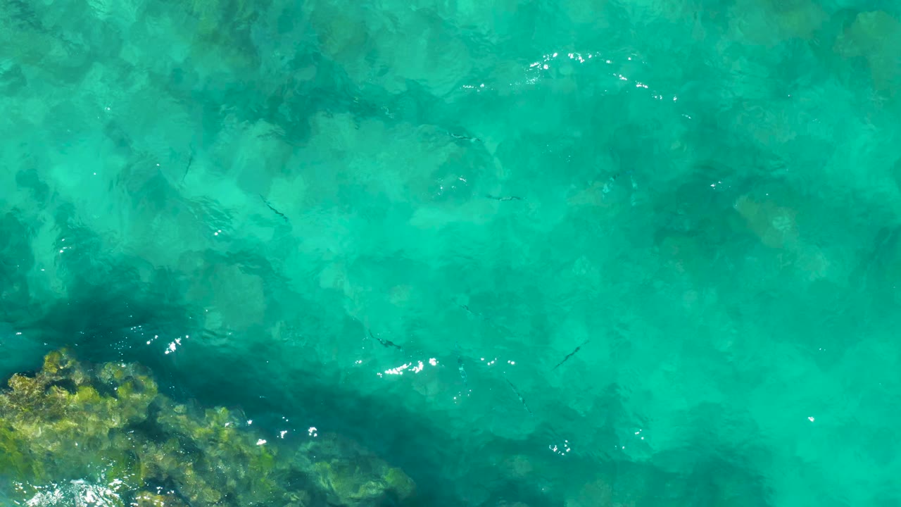 Overhead frame of clear turquoise water in East Cape BCS Mexico, with darker submerged reef texture near the lower left and fish shapes scattered under the rippled surface, no visible people.