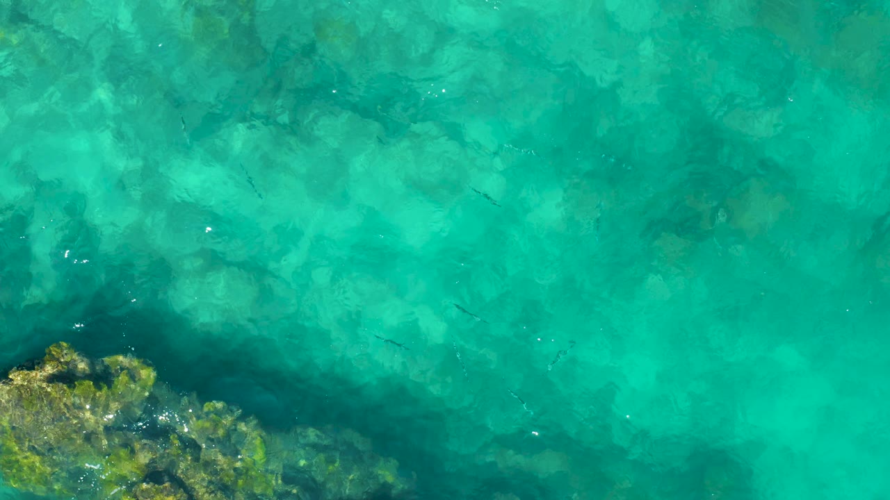 Clear turquoise ocean in East Cape BCS Mexico seen from above, with a reef edge along the lower left, darker submerged patches through the center, and no visible people.