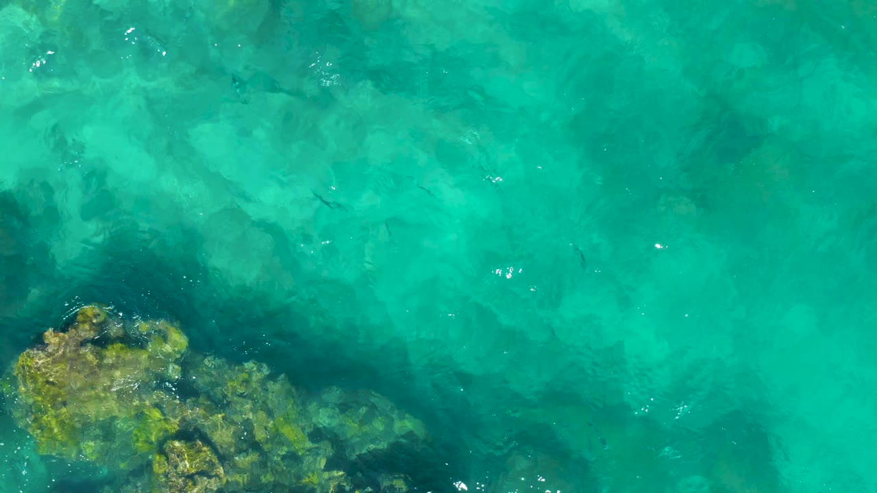 Aerial view of turquoise East Cape water with a larger submerged rock cluster near the lower edge and no visible people.