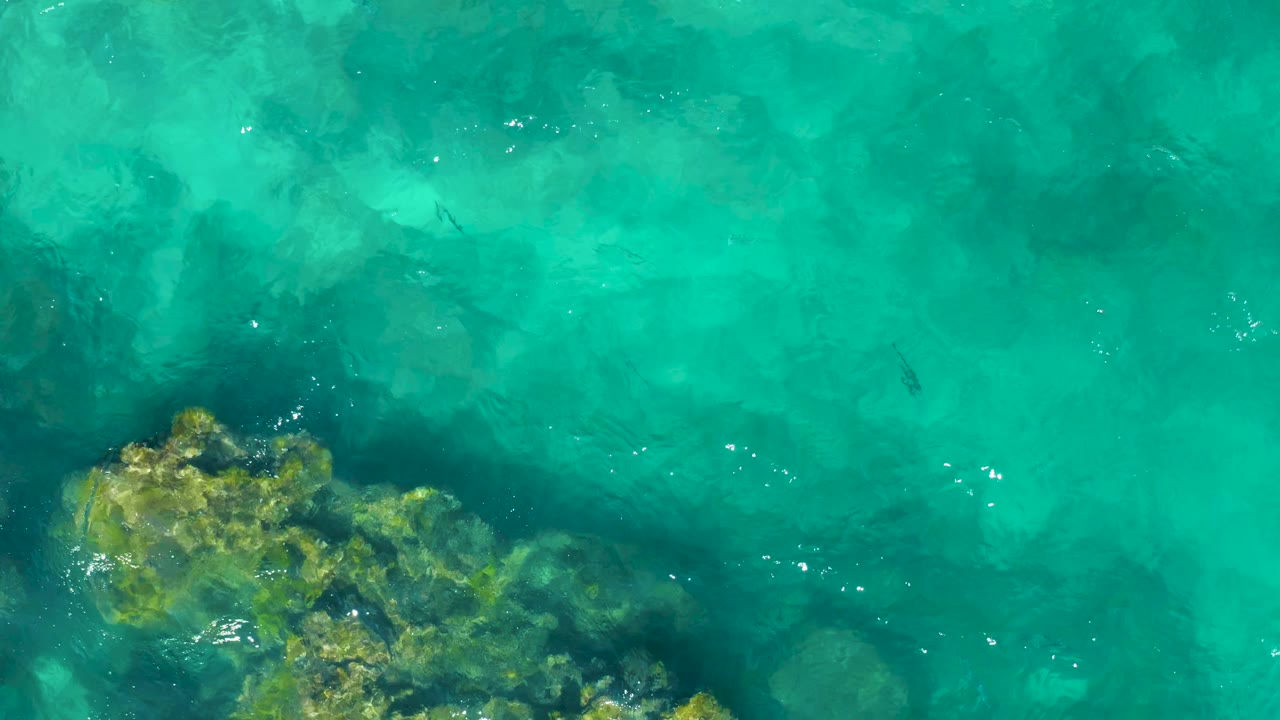 Turquoise ocean water reveals a darker rocky patch near the lower frame in East Cape, Baja California Sur, with no visible people.