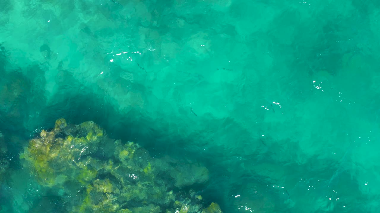 A straight-down aerial frame shows turquoise water over clustered underwater rocks in East Cape, Baja California Sur, with no visible people.