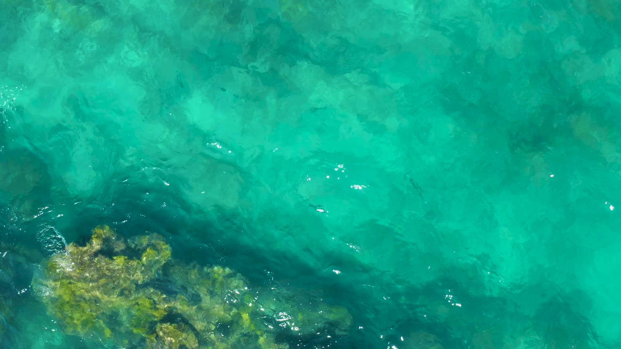 Aerial-looking frame over clear turquoise water in East Cape BCS Mexico, with darker seabed shapes drifting across the lower half and no visible people.