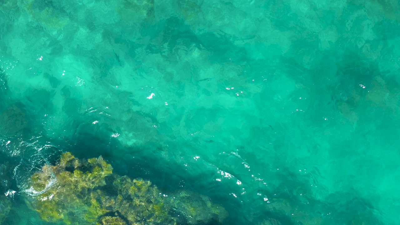 Top-down East Cape BCS Mexico water view with a larger rocky seabed patch along the lower-left edge, bright reflections, and no visible people.