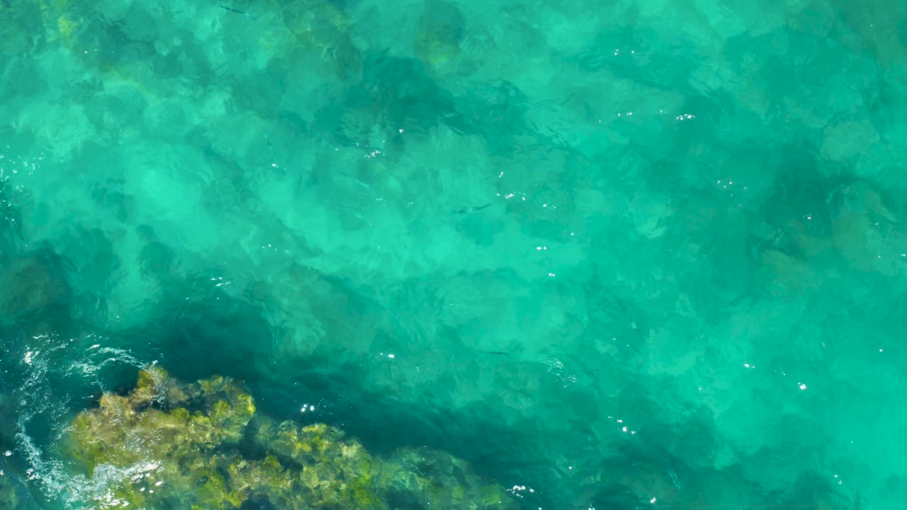 Clear turquoise water in East Cape BCS Mexico with a submerged rocky patch entering from the lower-left corner, faint wave texture, and no visible people.