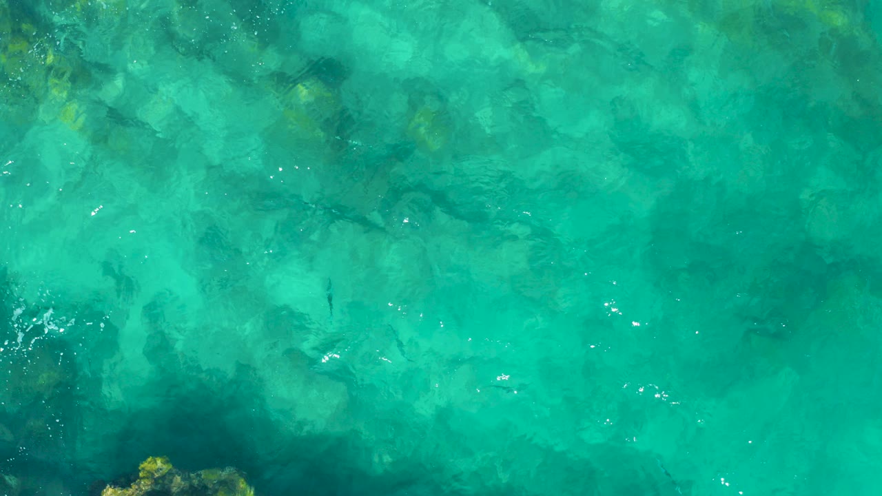 Top-down view of turquoise water over a rocky seabed in East Cape, Baja California Sur, Mexico, with faint ripples and no visible people.
