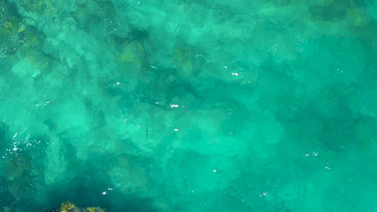 Shallow turquoise ocean water in East Cape, Baja California Sur, Mexico, with scattered sun flecks and soft rock patterns visible below, with no visible people.
