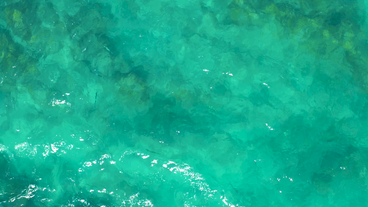 Top-down view of clear turquoise ocean water in East Cape, Baja California Sur, with dark underwater rock shapes and no visible people.