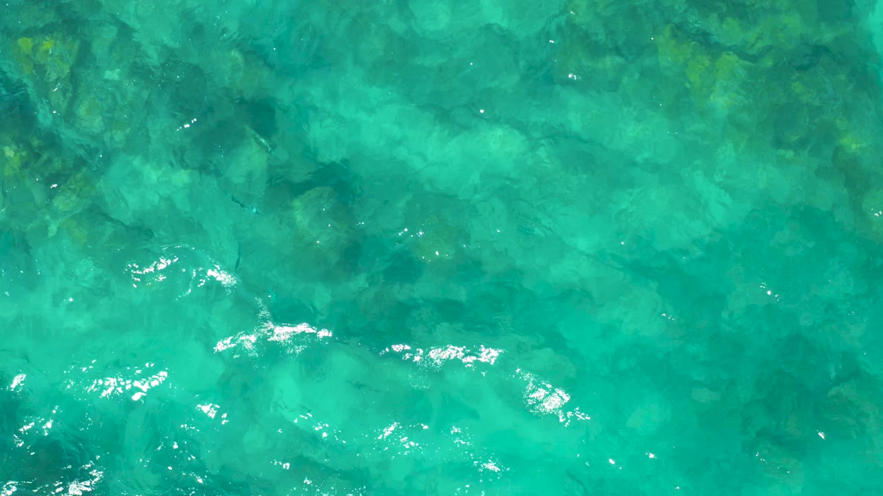 Turquoise shallow ocean water shows brighter white ripple highlights over mottled underwater rock forms, with no visible people.
