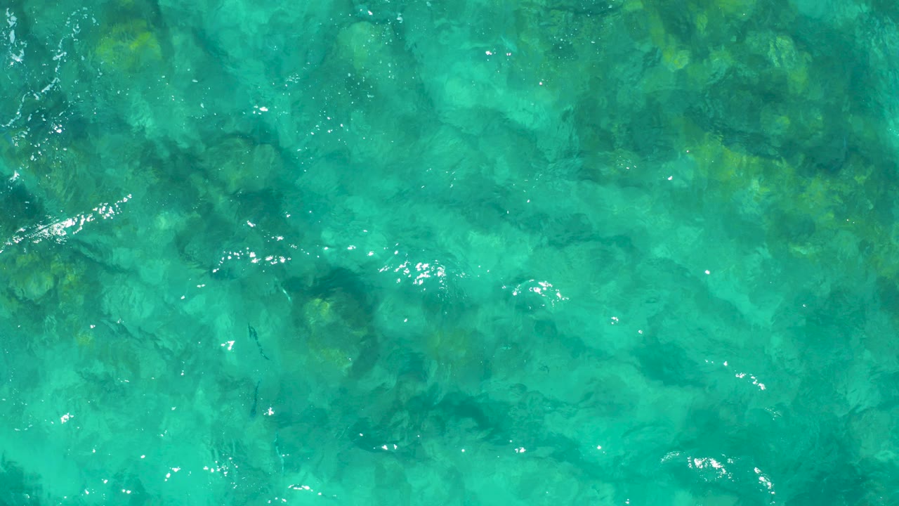 Overhead view of clear turquoise water in East Cape BCS Mexico, with dark submerged rocks or coral visible beneath light ripples and no visible people.