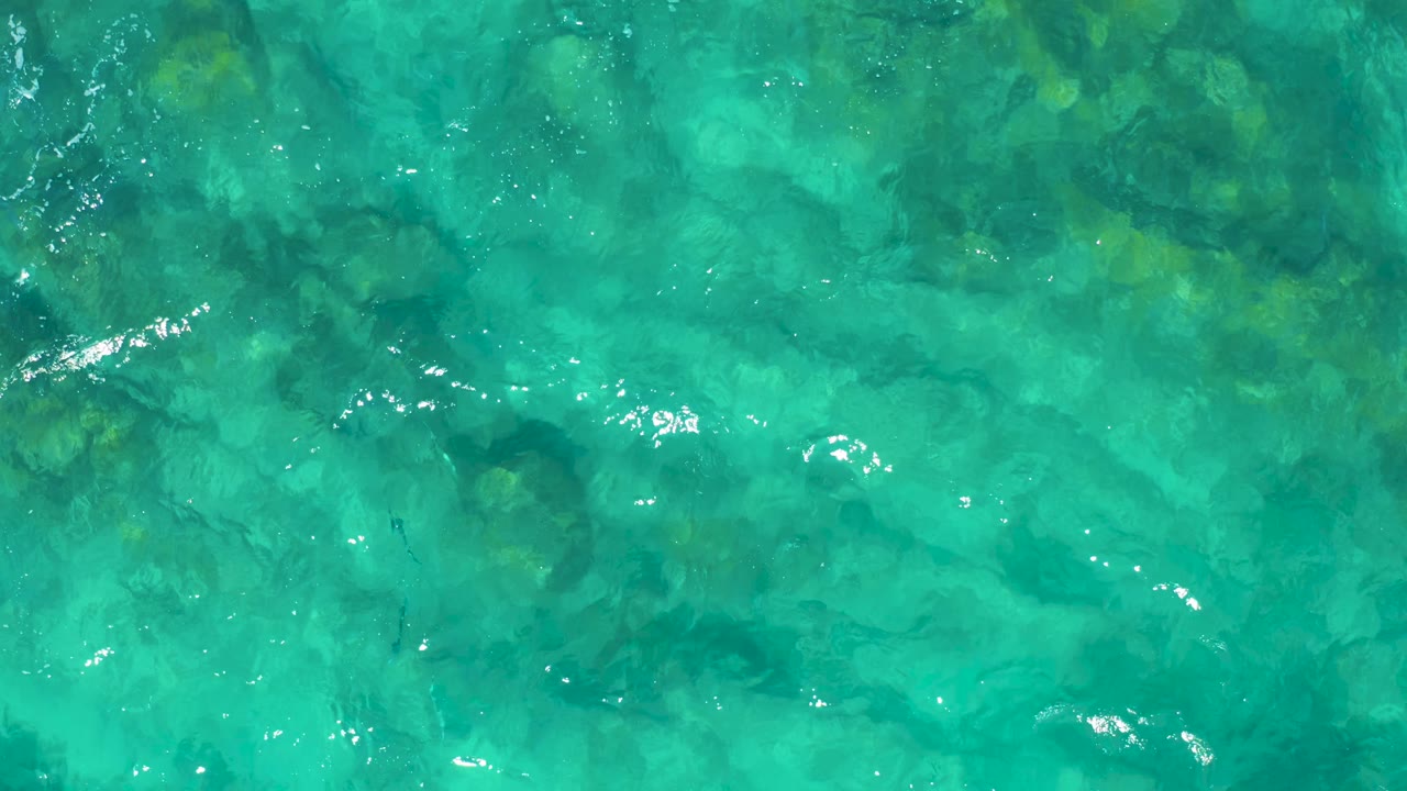 Clear turquoise ocean water reveals darker rocky patches below the surface, with small bright ripple highlights and no visible people.