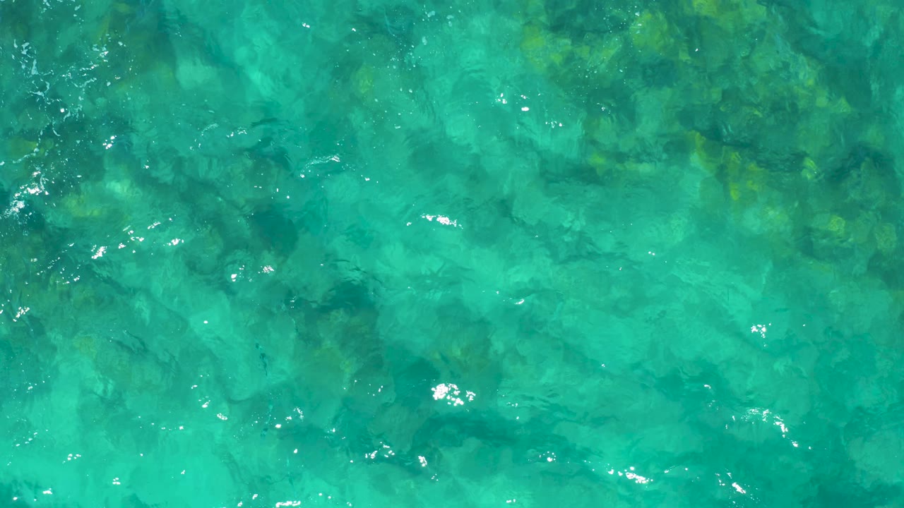 A full-frame overhead view of shallow turquoise sea shows rocks or coral under the surface, with scattered white glints on the moving water and no visible people.